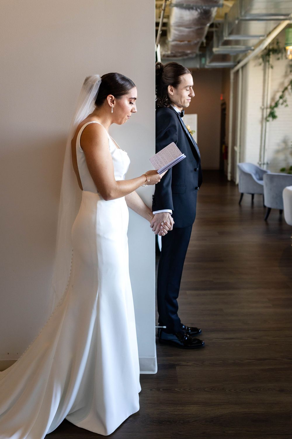 A couple in formal wedding attire holding hands while standing apart, reading letters in an indoor venue.