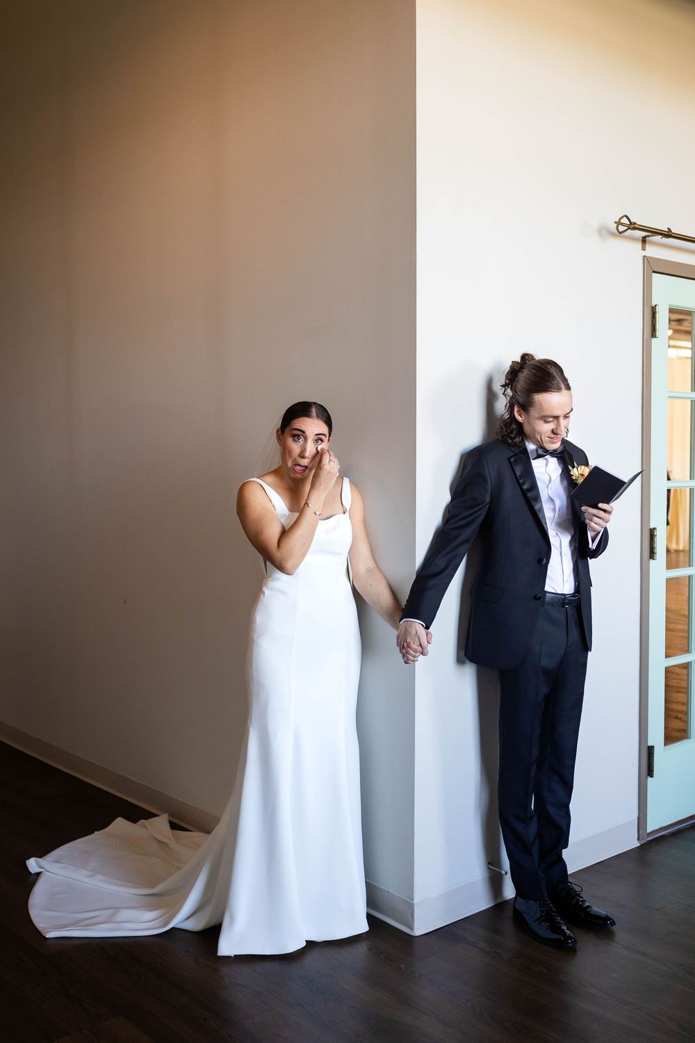 Bride and groom hold hands while standing on opposite sides of a wall in a sweet first look moment.