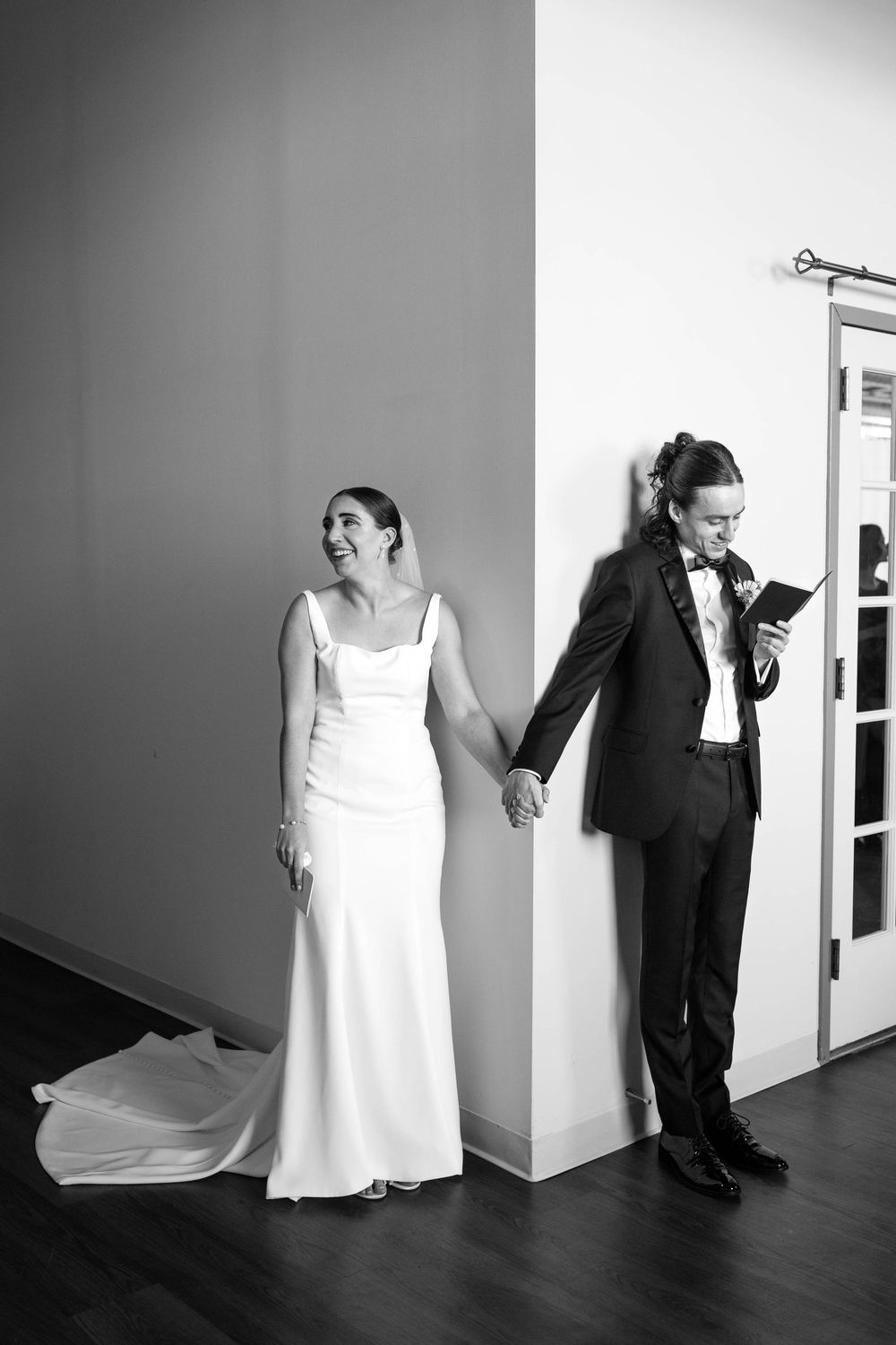A wedding couple holds hands while standing on opposite sides of a corner wall in black and white.