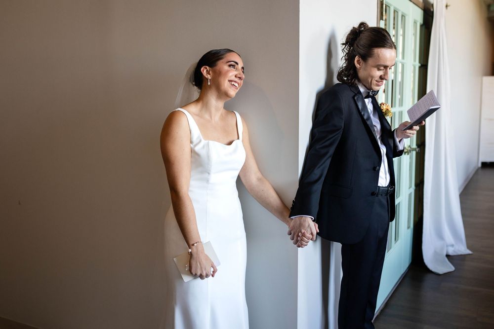 A couple in wedding attire hold hands while standing apart, separated by a wall during their first look moment.
