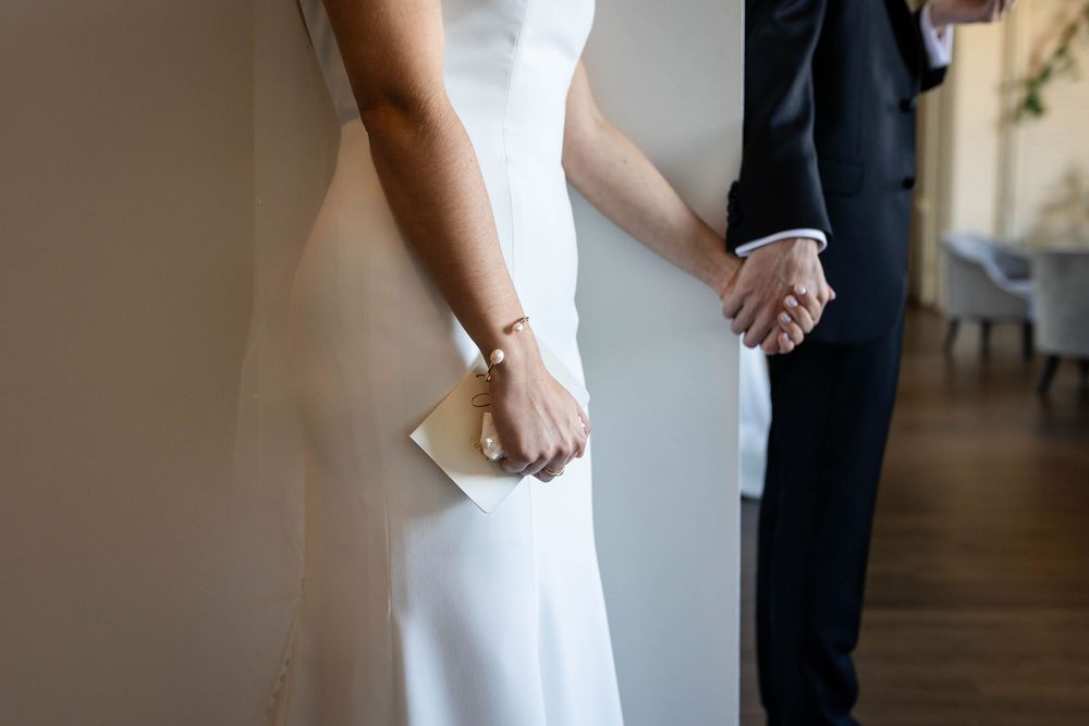 A couple holds hands while standing indoors, with the bride in a white dress holding a small clutch purse.