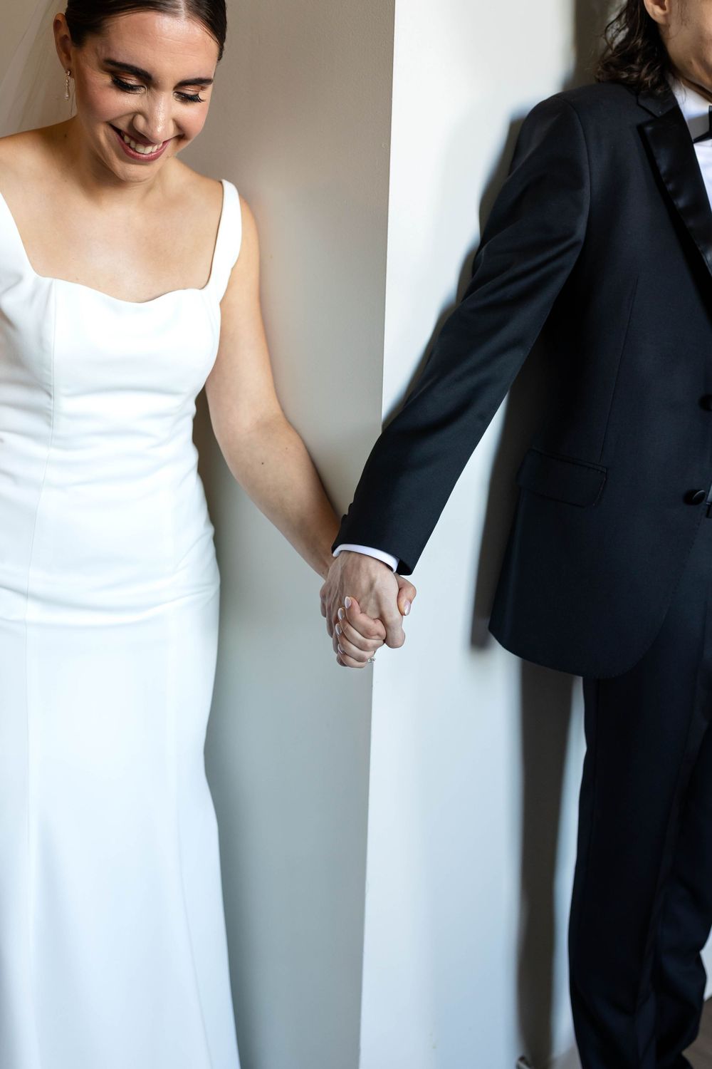 A bride in a sleek white gown holds hands with her partner in a classic black tuxedo against a neutral wall.