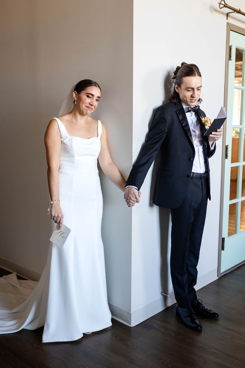 A couple shares a playful moment during their first look, separated by a wall while holding hands.