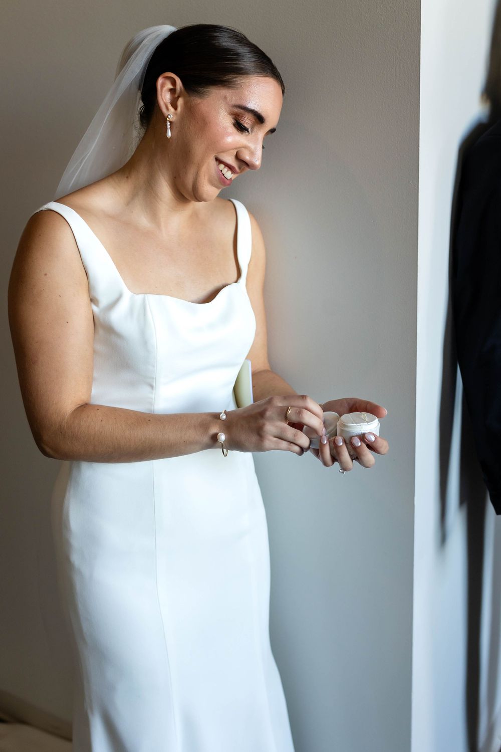 A bride in a sleek white dress admires her wedding ring near a window.