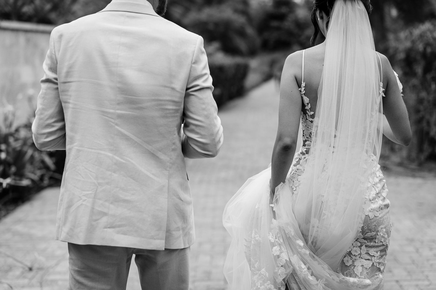 A couple holds hands while walking through a tropical garden with palm trees and ornamental grasses.