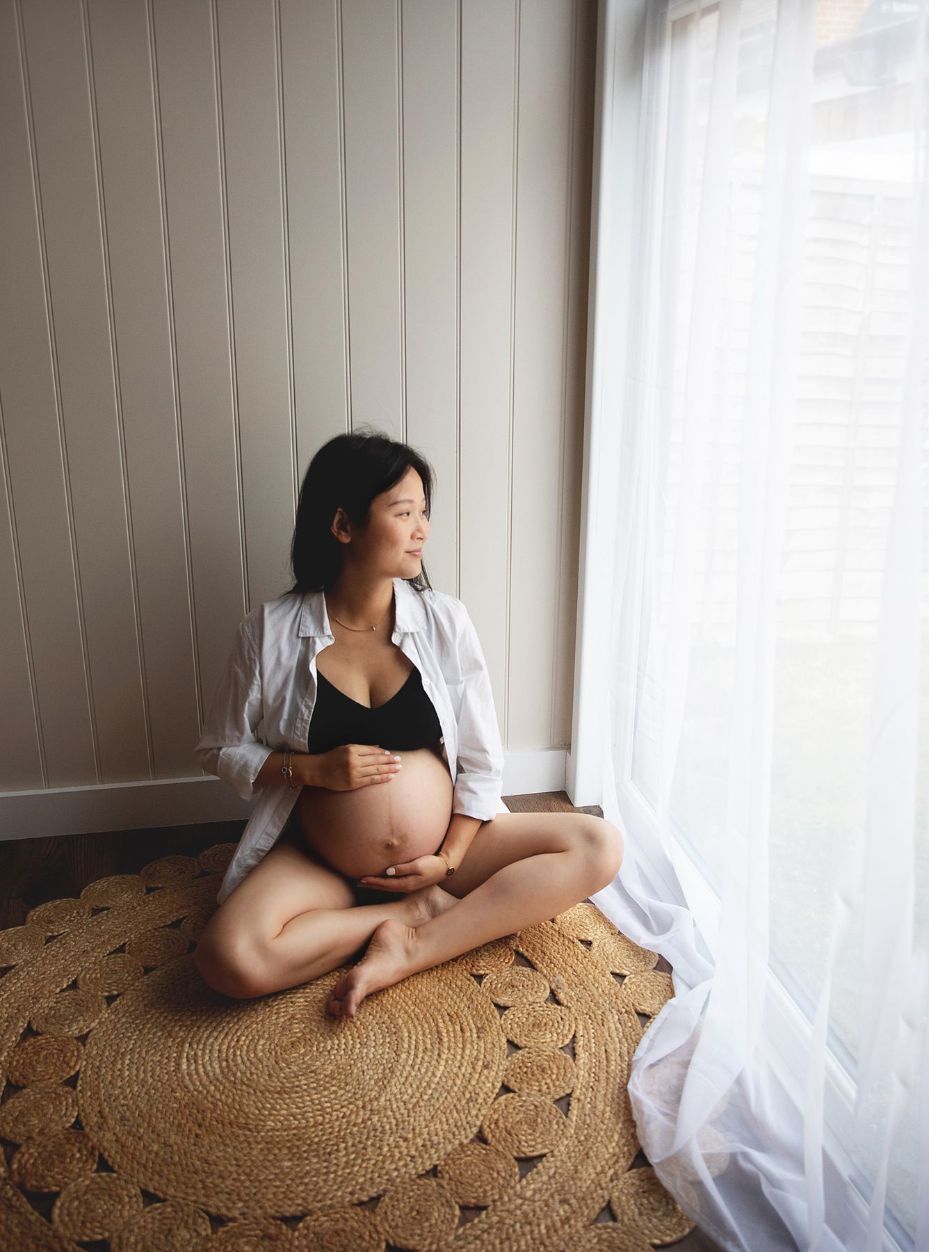 Expectant mother in white shirt sits cross-legged on round jute rug by window during maternity photoshoot. During a maternity photoshoot