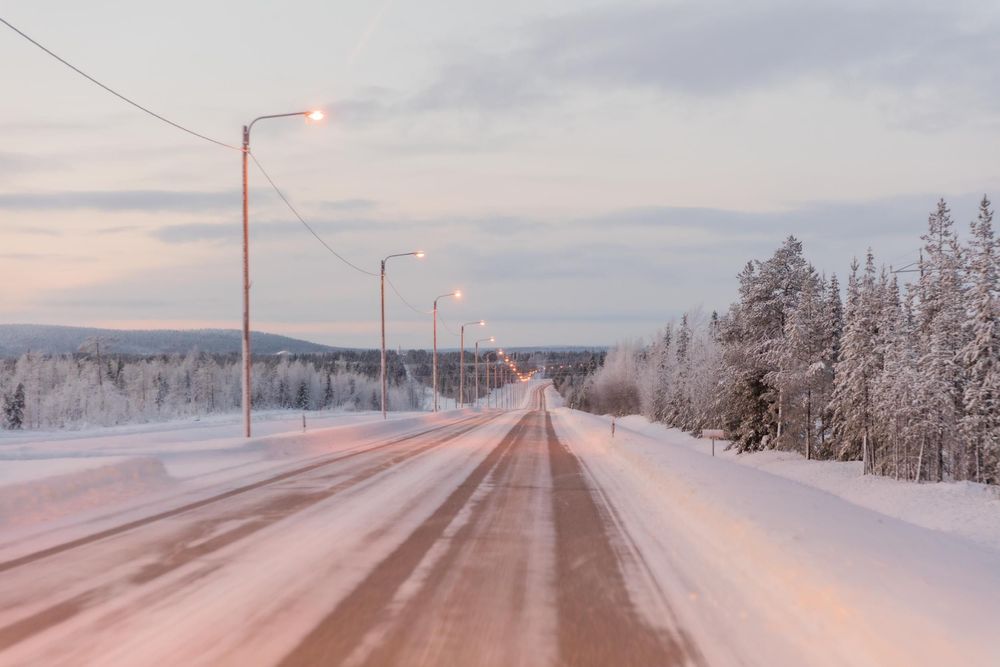 A snow-covered rural road stretches into the distance with illuminated lampposts and frost-covered trees in winter twilight.