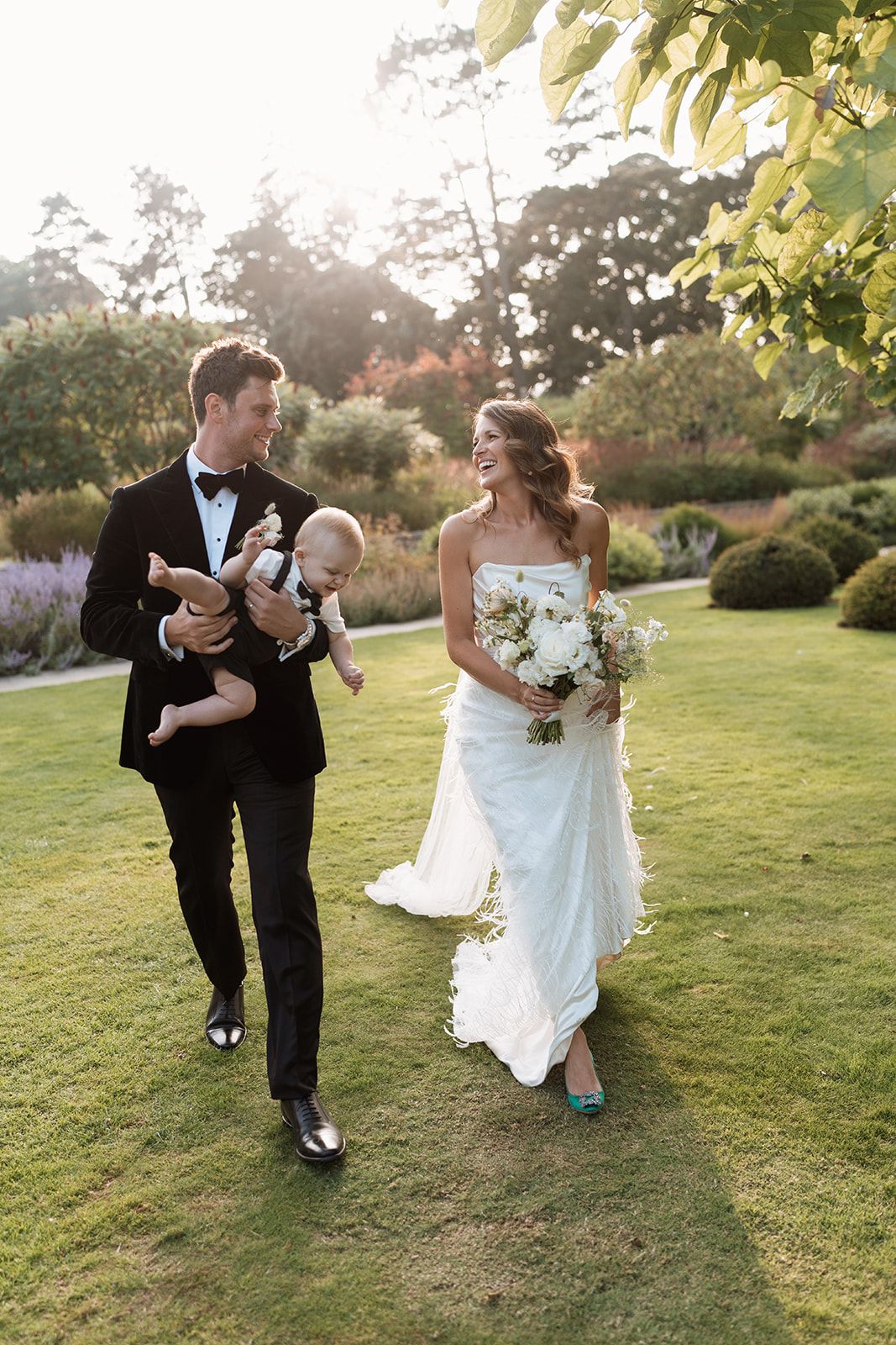 Newlyweds walk together on a lush garden lawn at sunset, with bride in white dress holding flowers.