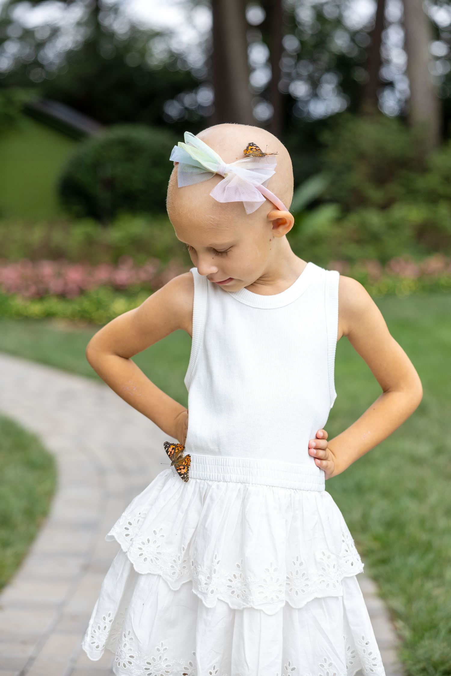 A small child in a white eyelet lace dress and hair bow stands on a stone pathway in a garden setting.