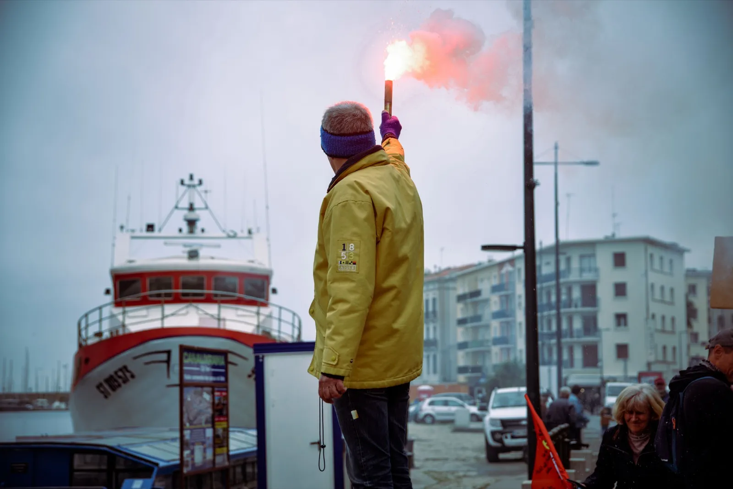 Homme en ciré jaune brandissant un fumigène rouge face à un bateau amarré, dans une ambiance de protestation portuaire.