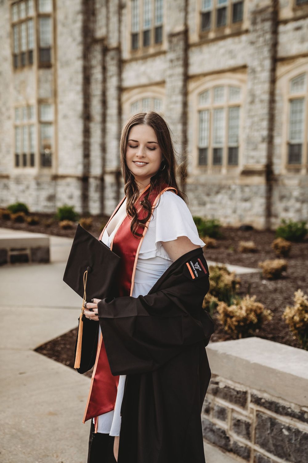 Young woman in black graduation gown and red sash holds cap outside stone university building.