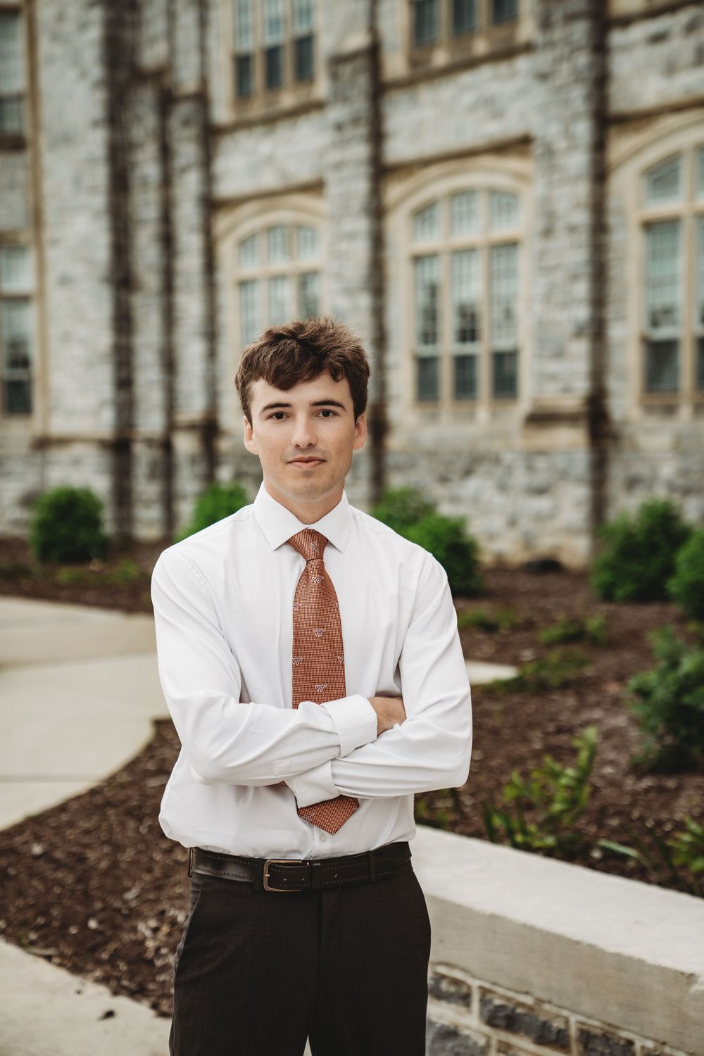 Young man in white shirt and orange tie stands arms crossed before a stone university building.
