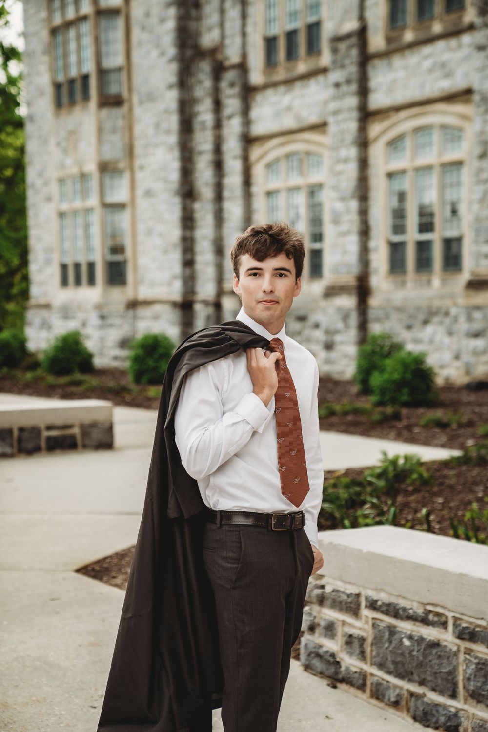 Graduate in white shirt and tie holds dark academic robe over shoulder outside stone building.