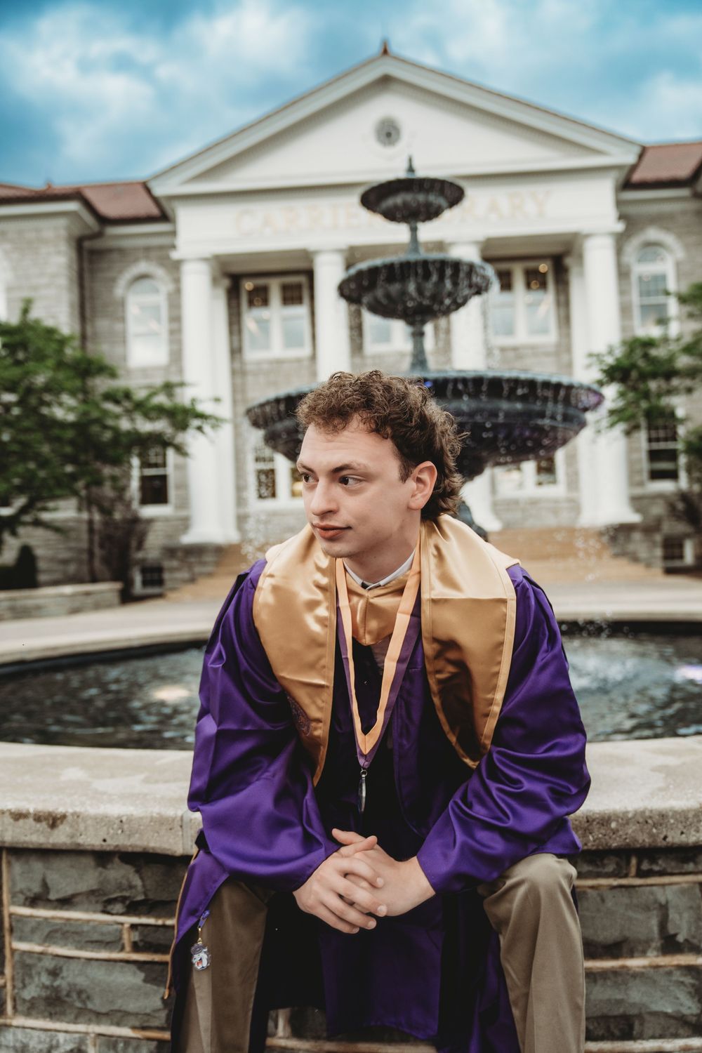 Young man in purple and gold graduation regalia sits by a fountain outside Carrier Library on a cloudy day.