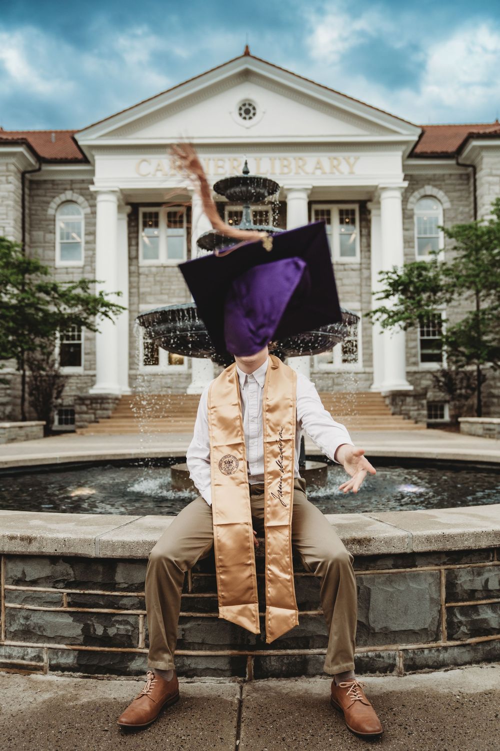 Graduate tosses purple cap in front of campus library fountain, wearing gold honor stole and white dress shirt.