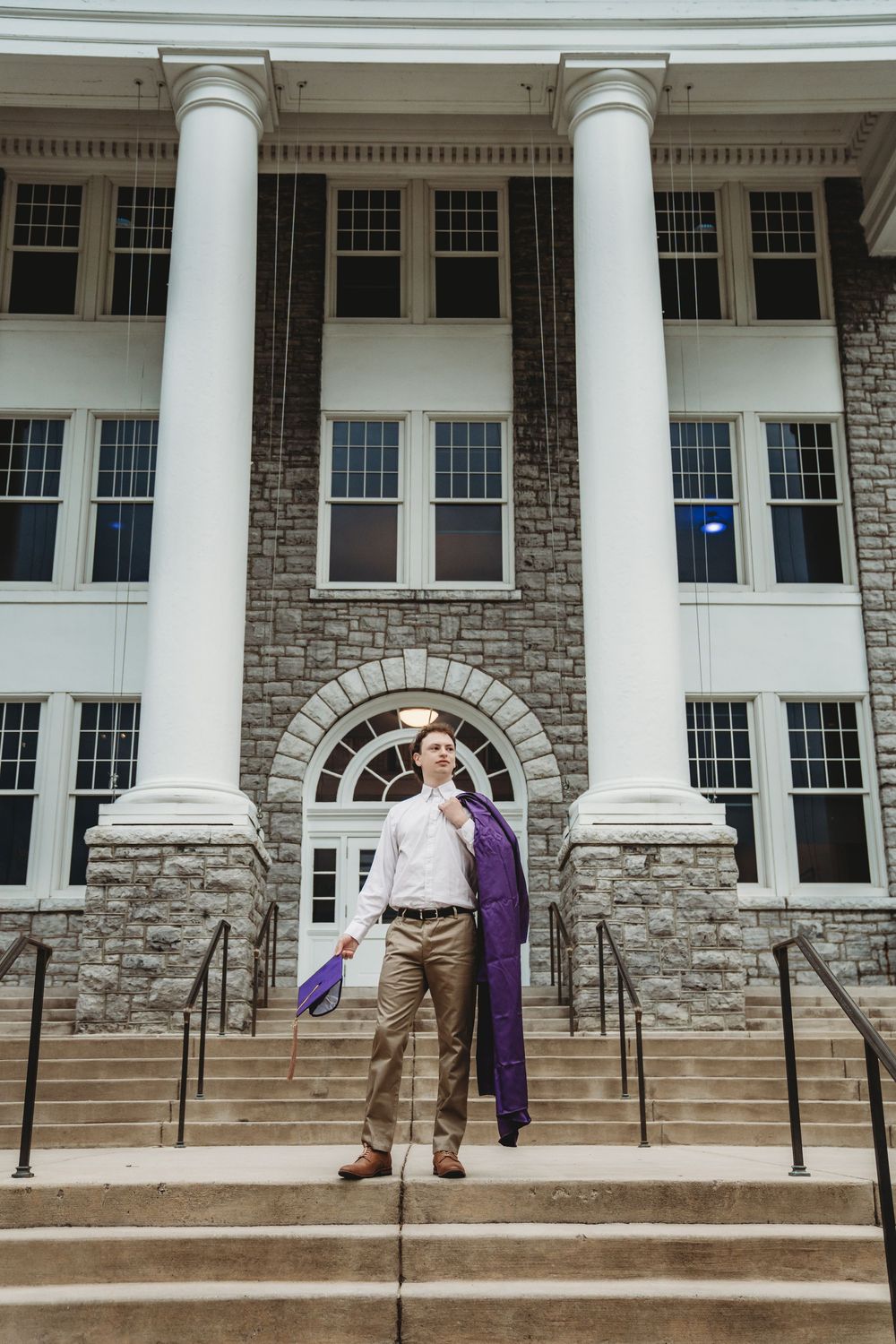 Young man in white shirt and purple jacket poses on steps of a stone building with white columns.