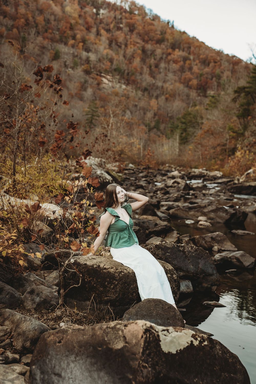 A person in a white dress reclines on dark rocks near a calm stream in an autumn mountain setting.