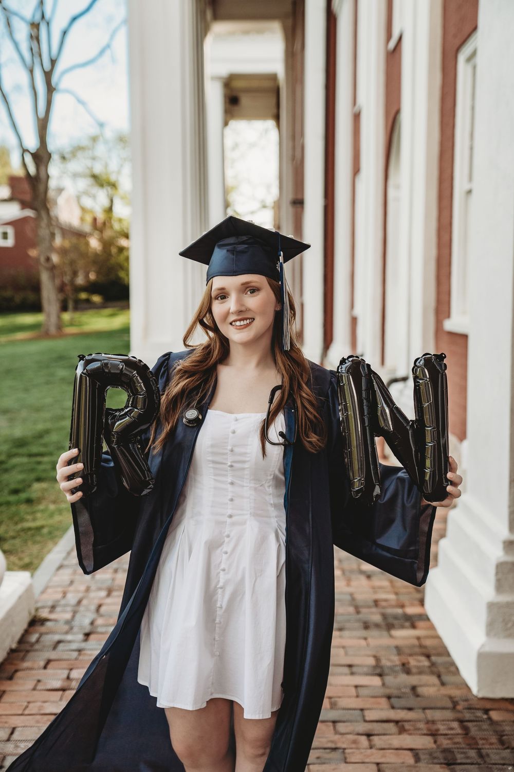 A smiling graduate in cap and gown holds black letter balloons spelling RN on a brick pathway between white columns.