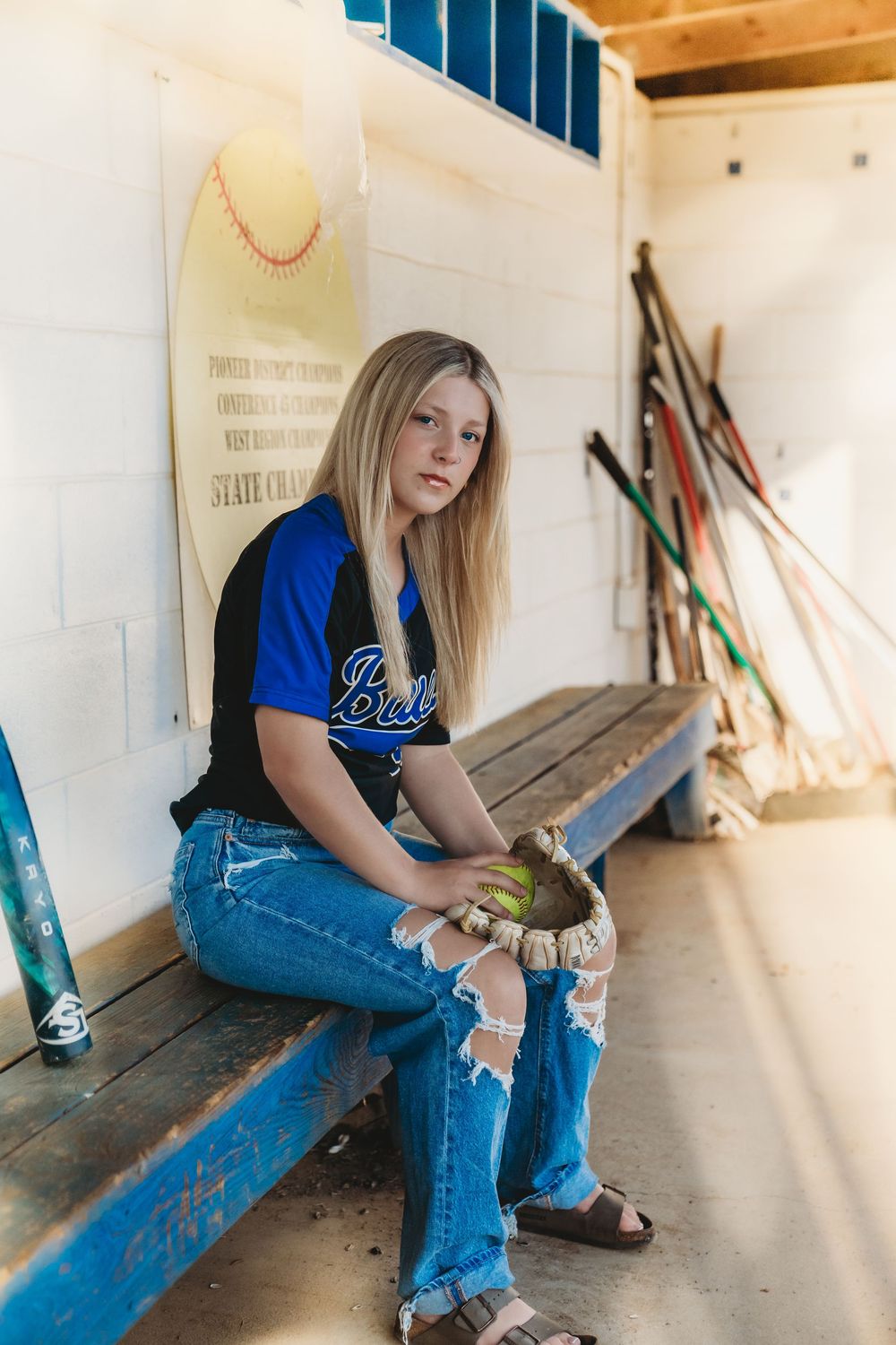 Casual senior photos in dugout wearing blue jeans and softball gear.