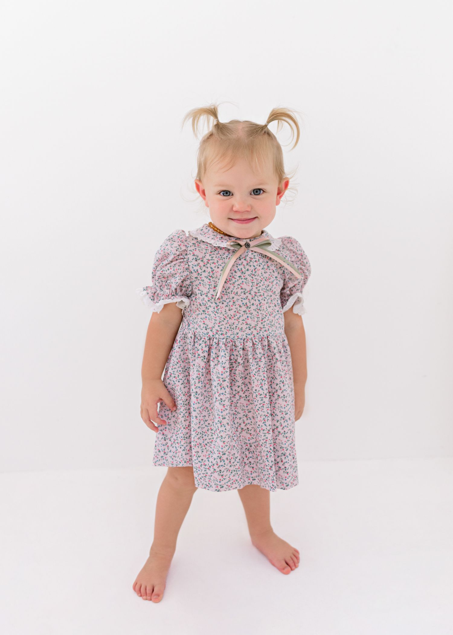 Sweet toddler in floral print dress with pigtails stands barefoot against white background, smiling brightly.
