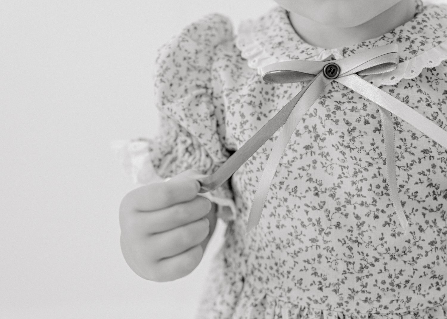 Close-up black and white photo of child's hands holding scissors near a floral print dress with ribbon bow detail.