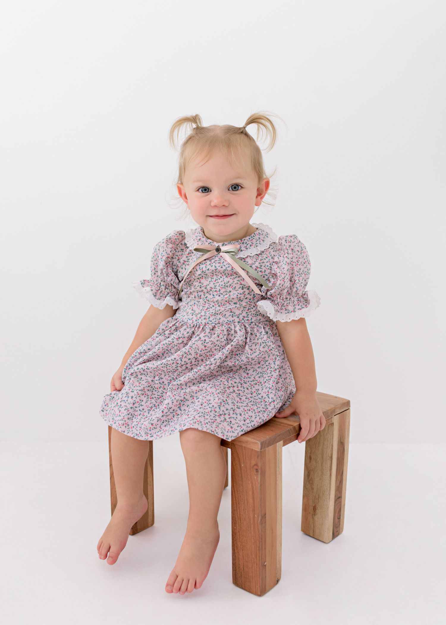 A toddler in a floral vintage-style dress with puff sleeves sits on a wooden stool against a white background.