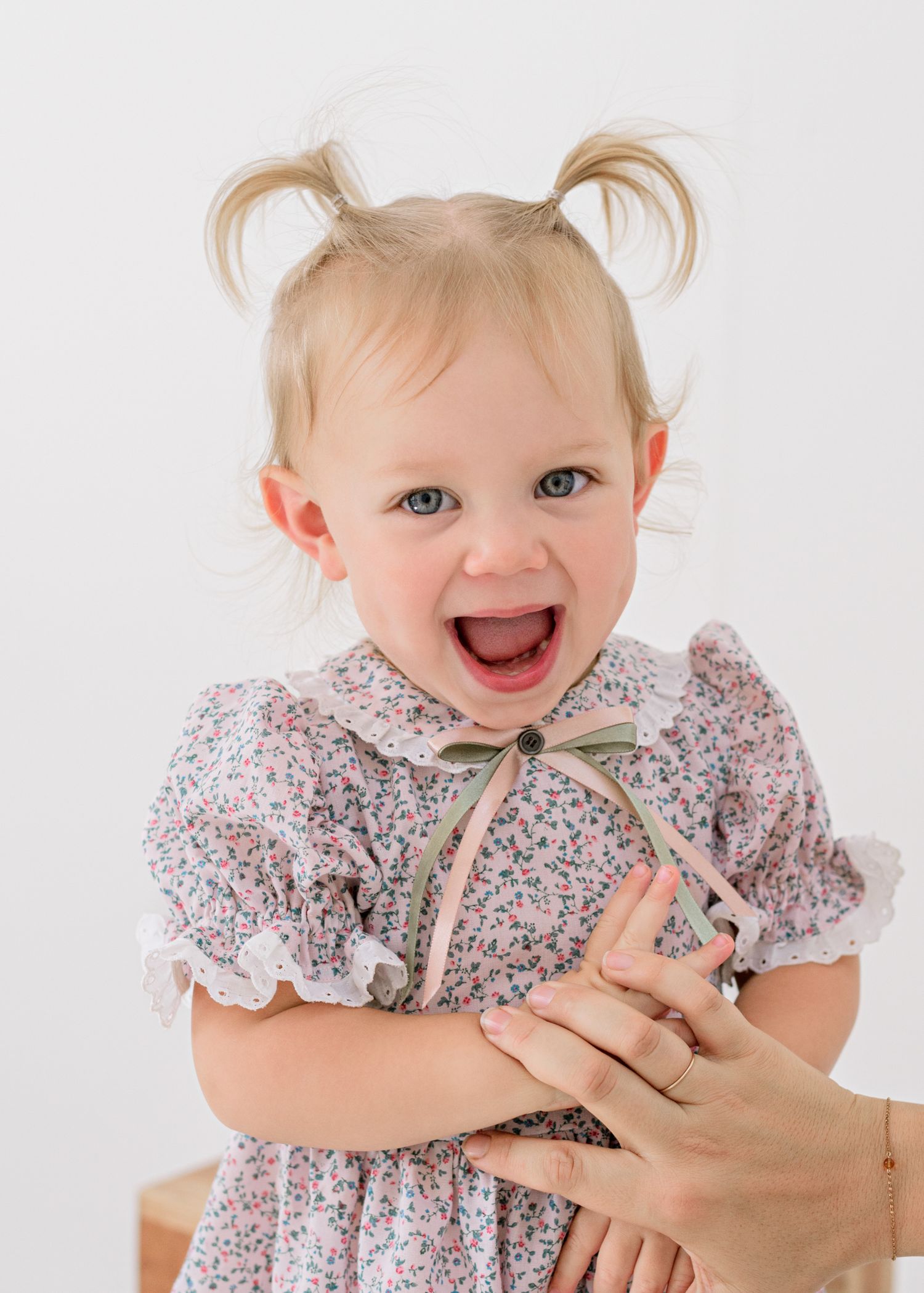 Joyful toddler wearing floral dress with puff sleeves and bow laughs against white background.