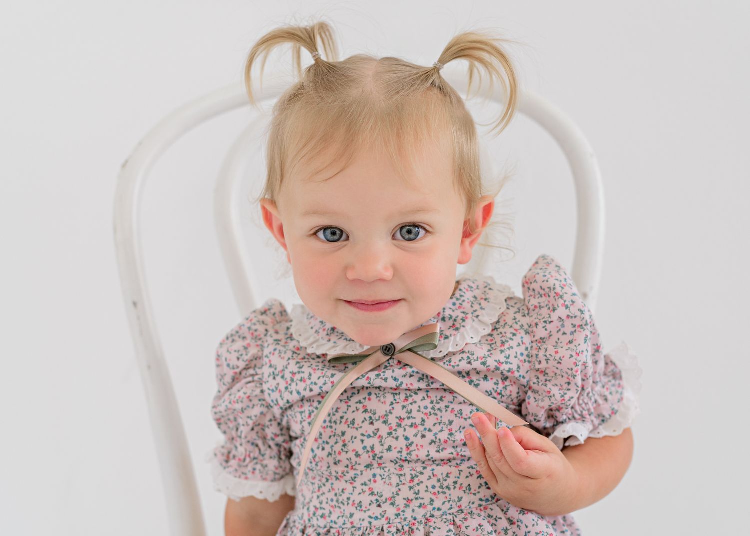A small child in a floral dress with blonde pigtails sits on a white chair.