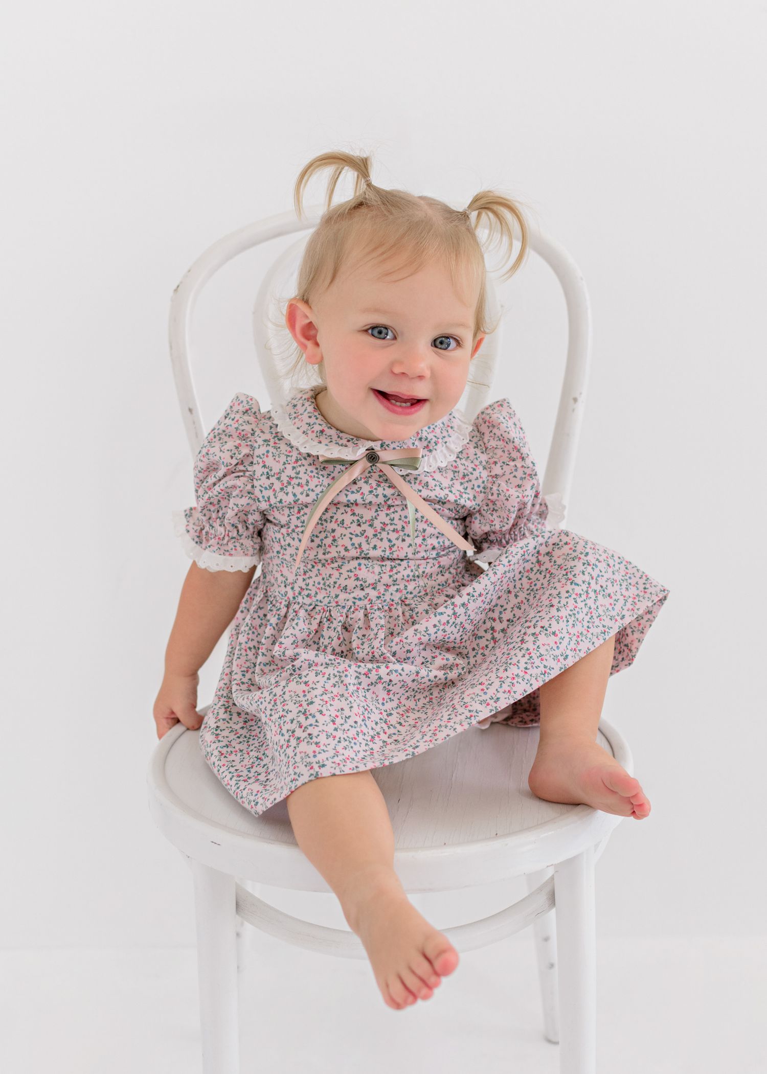 Toddler wearing floral print dress sits smiling on white wooden chair with pigtails.