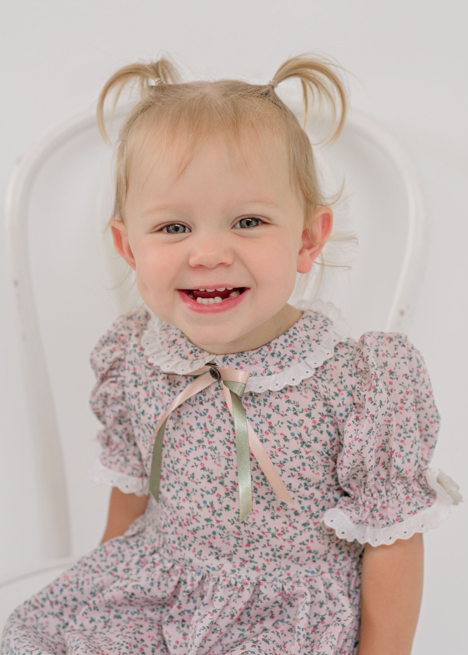 Portrait of a cheerful toddler wearing a floral vintage dress with lace collar and pigtails against white backdrop.