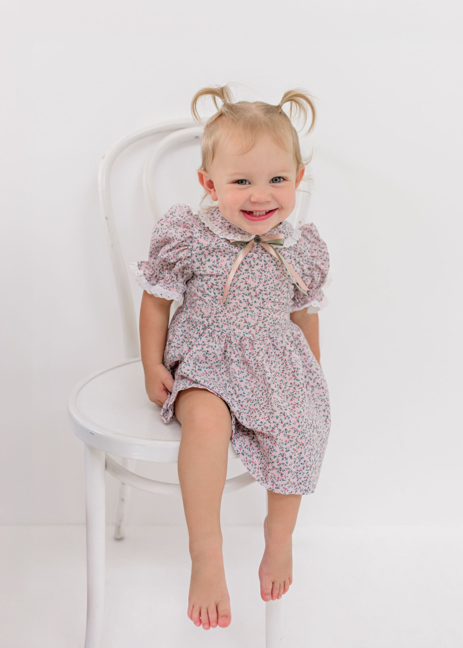 Small child in vintage-style floral dress perched happily on white chair.