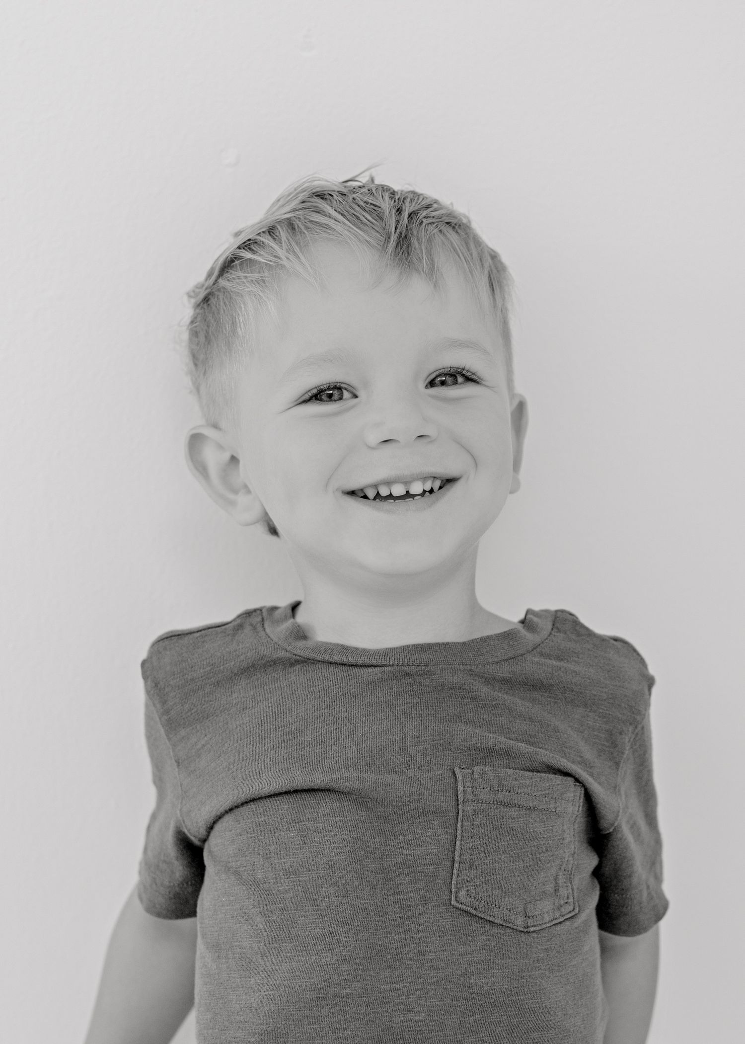 Black and white portrait of a child wearing a t-shirt with pocket smiling brightly against a plain background.