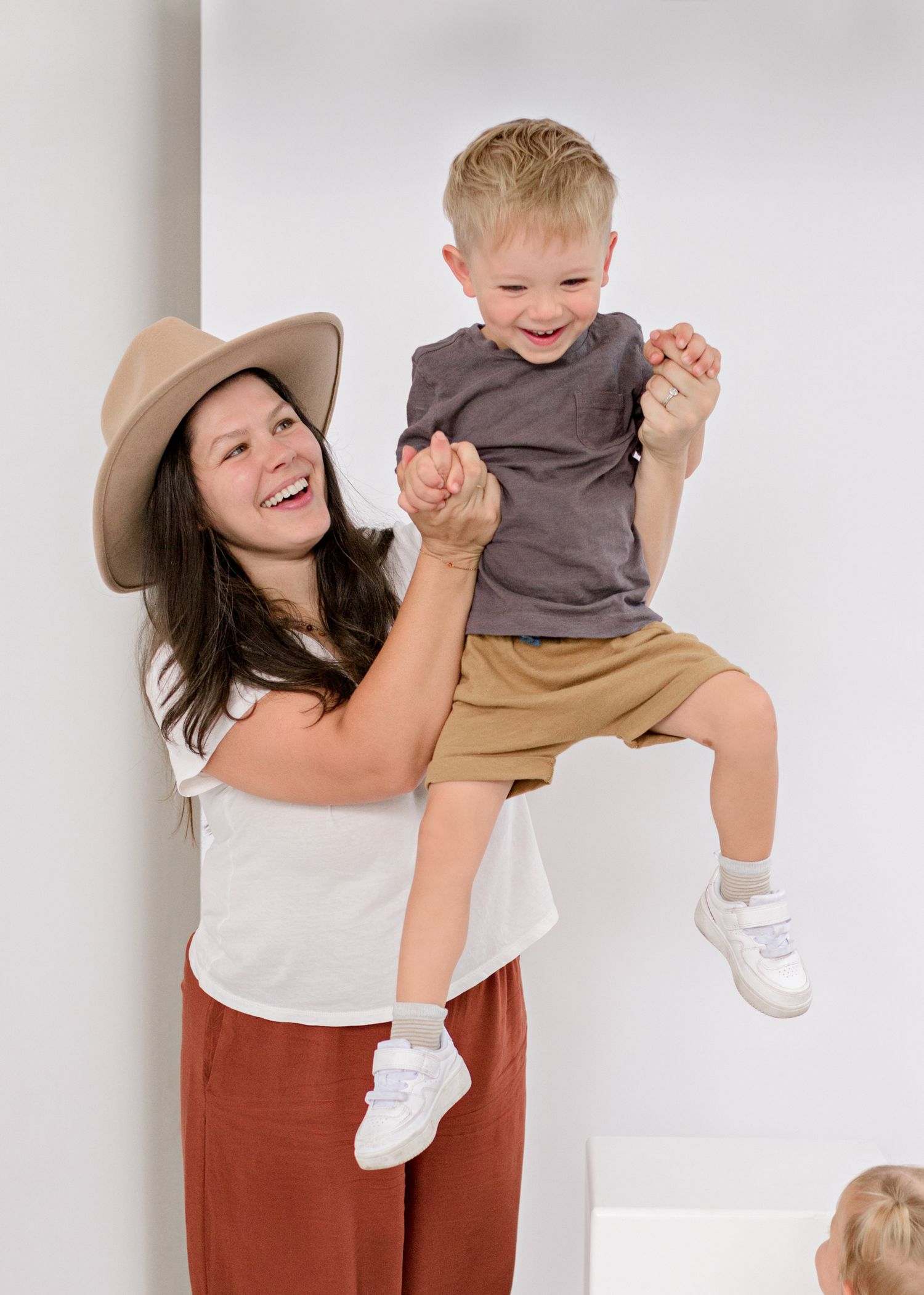 A joyful moment captured as a woman in a hat lifts a small child wearing grey shirt and khaki shorts against a white wall.