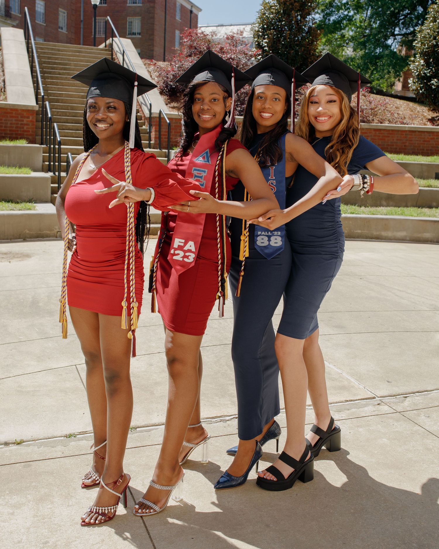 Four graduates in caps and gowns pose together on campus steps wearing red and navy dresses.
