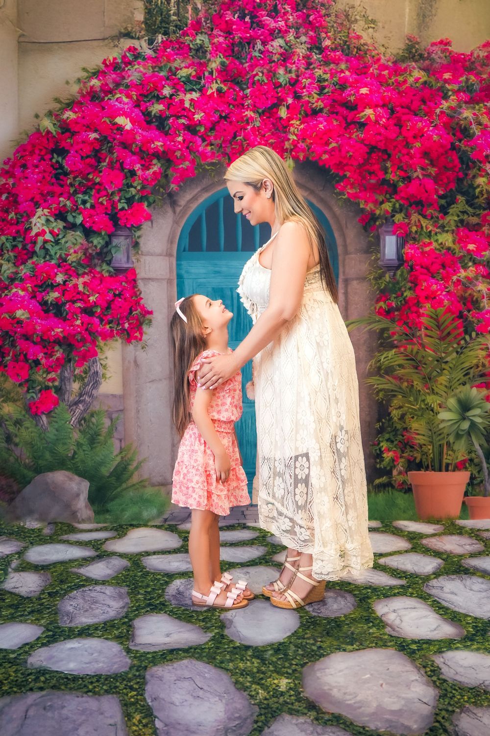A parent and child share a tender moment under an archway draped with vibrant pink bougainvillea on a stone pathway.