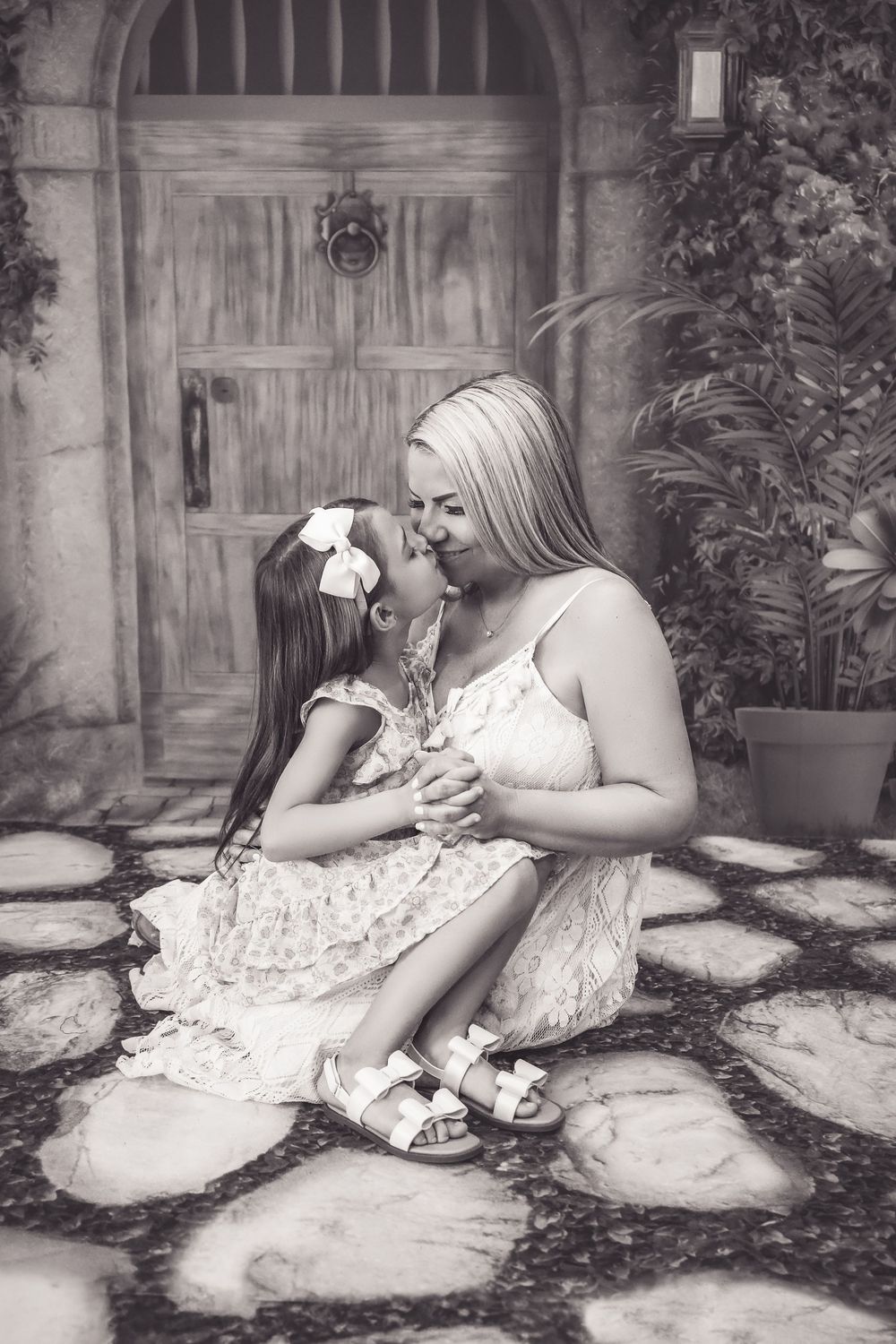 A mother and daughter in matching white lace dresses sit together on a stone patio in black and white photography.