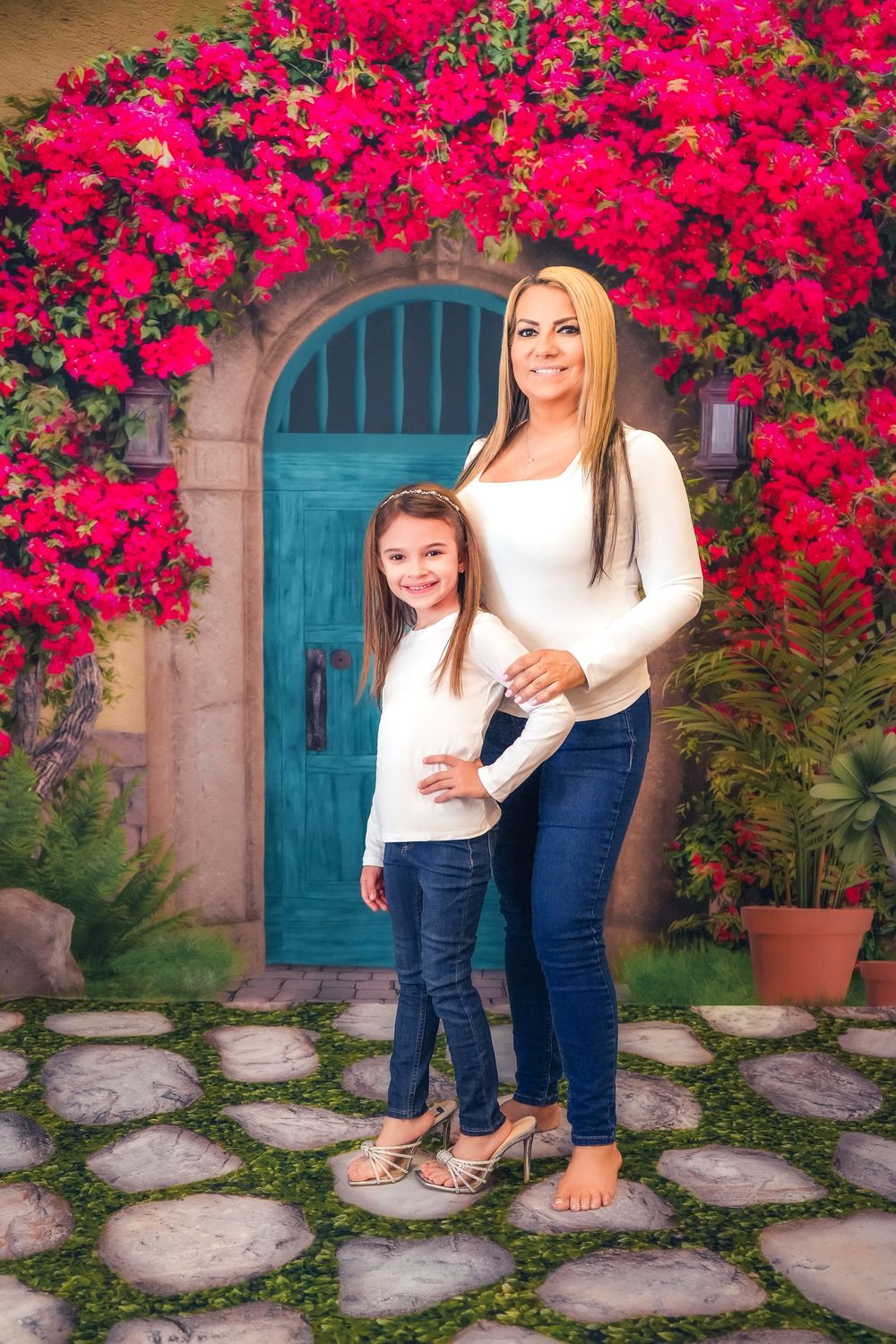 Two people in matching white sweaters and jeans pose in front of a turquoise door with vibrant pink bougainvillea flowers.