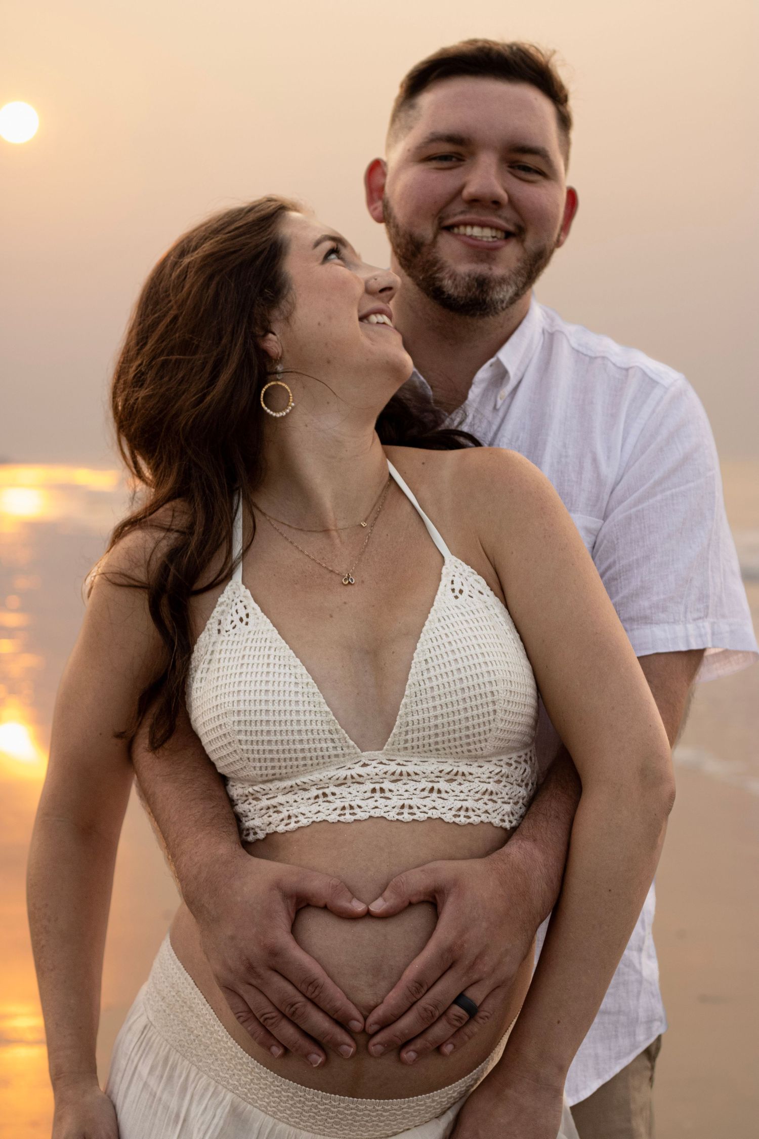 Expected parents on the beach at sunrise with husband holding mothers belly and she is looking at him by Raley and Roots Photography.