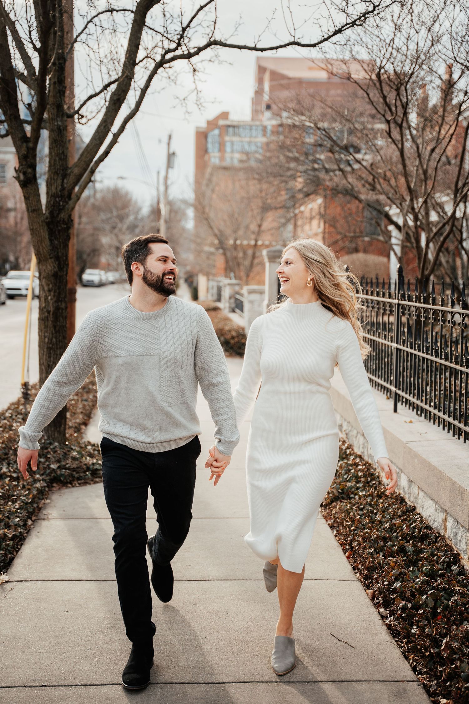 Couple in matching cream sweaters walk hand in hand down a city sidewalk on an autumn day.