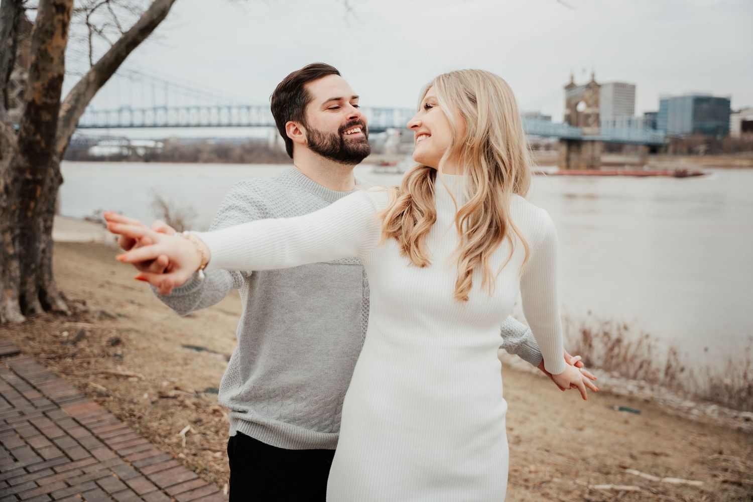 A couple laughs together while standing along a riverside path with a bridge visible in the background.