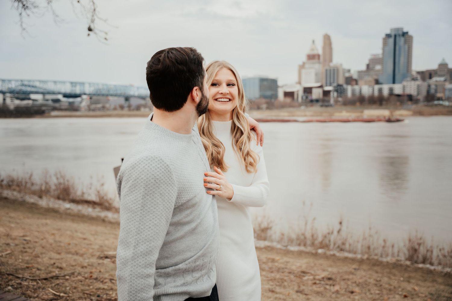 A couple shares a romantic moment along a riverside path with an urban cityscape in the background.