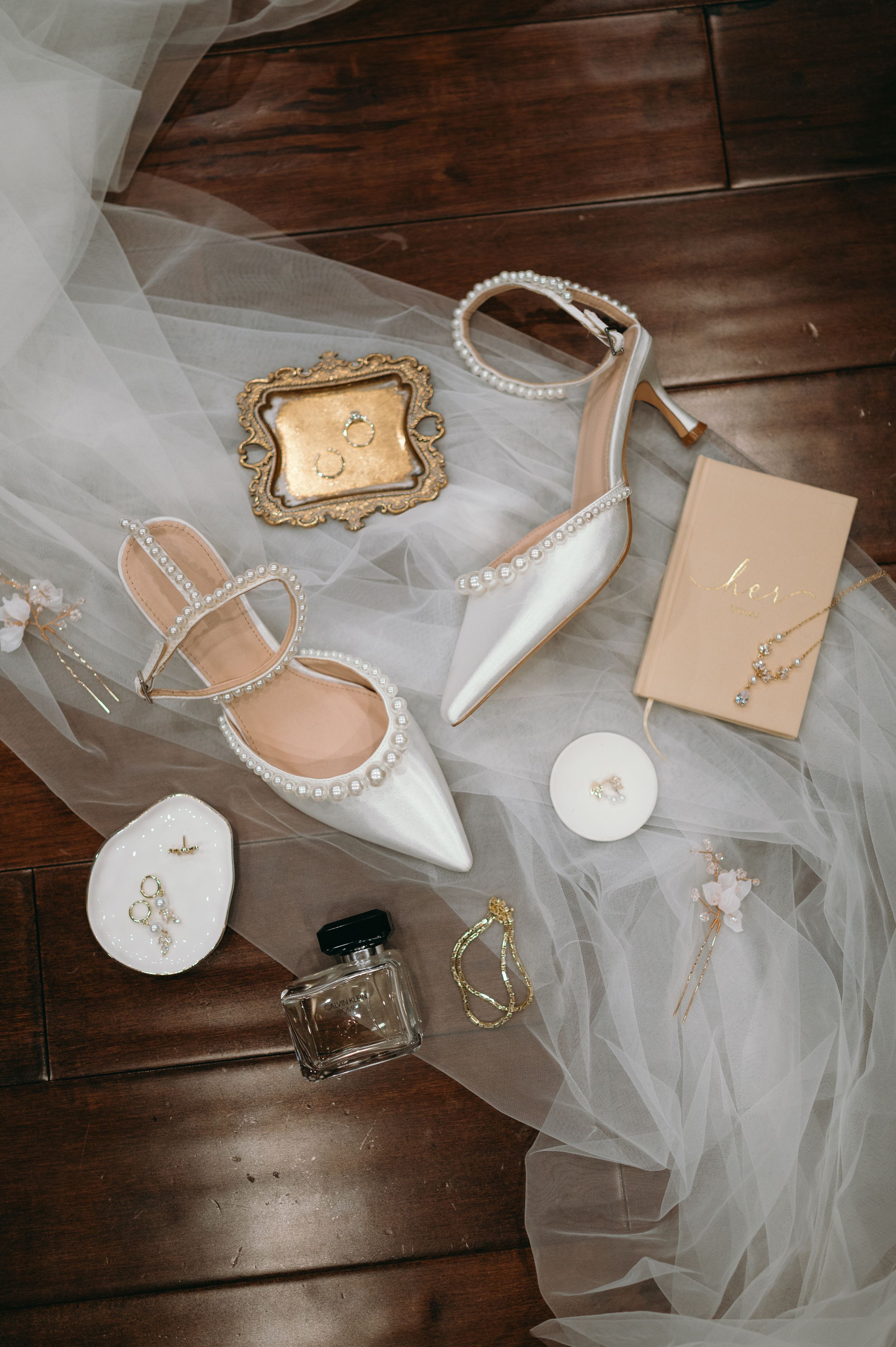 Wedding accessories including pearl-adorned shoes, perfume, veil and delicate details on wooden floor.