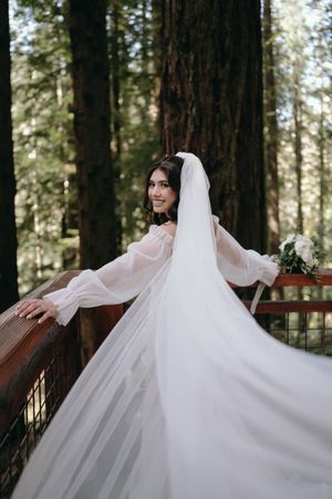 A bride in flowing sleeves and veil poses gracefully on a wooden deck in the forest.