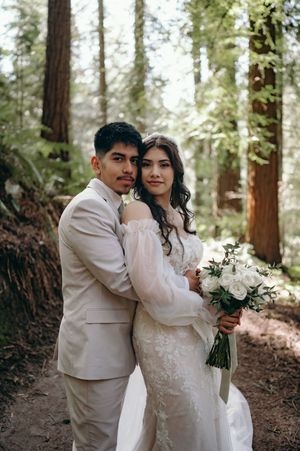 Couple poses in elegant wedding attire among towering redwood trees holding white bouquet.