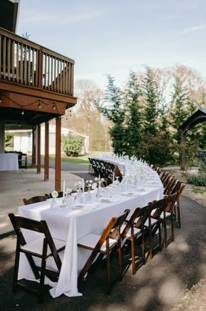 A long curved dining table with white linens and dark wooden folding chairs set up under a deck for an outdoor wedding reception.