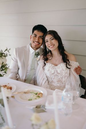 Newlywed couple in white attire smile while seated at their wedding reception table.