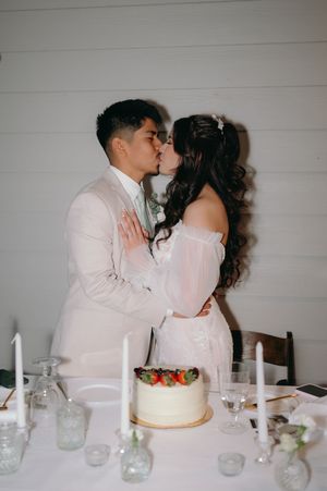 Couple shares romantic kiss behind their simple white wedding cake at reception.