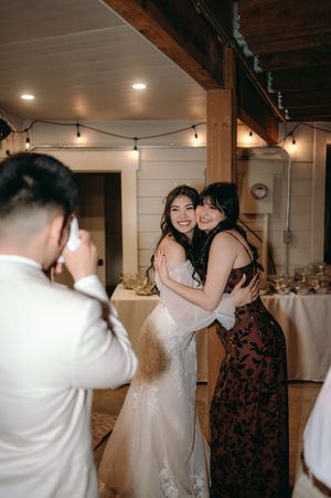 A wedding reception moment with a bride in white lace hugging a guest in a brown dress under string lights.