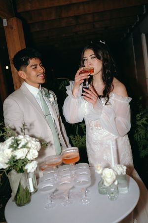Newlyweds share drinks at their wedding reception beside a table with champagne glasses and floral bouquets.