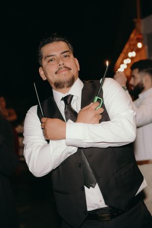 Event photographer in formal attire holding two camera flashes while posing at nighttime celebration.